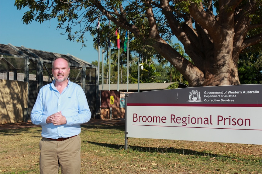 A grey-haired man in a blue shirt stands in front of a sign that reads "Broome Regional Prison".