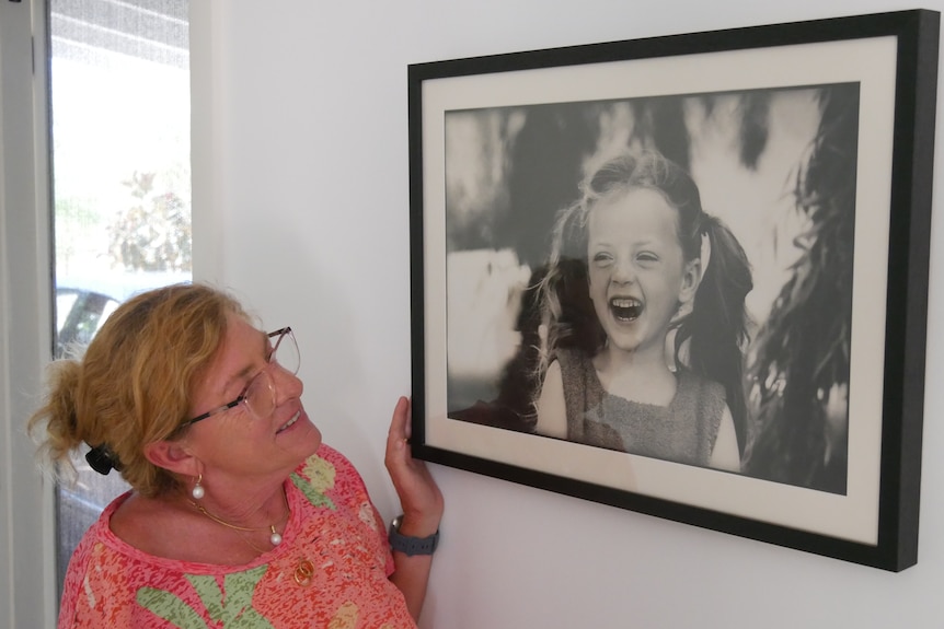 A woman in her fifties looks up nostalgically at a framed portrait of a young girl with pig tails laughing.