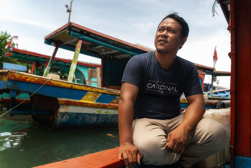 A young man wearing a blue Tshirt and beige pants sits on the edge of a boat in a marina, another boat behind him