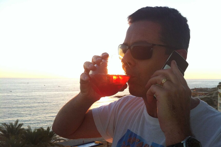 Matt Brown on phone and drinking drink with sunset over water in background.