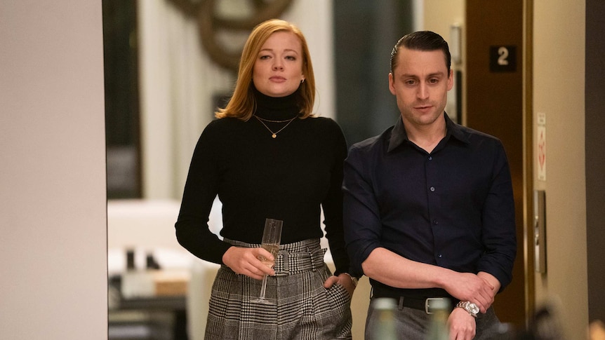 The two actors, both in dark expensive-looking clothing, stand in a doorway looking haughty. Snook holds a half-drunk champagne.