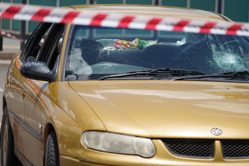 A gold car with a smashed windscreen, with crime tape in the foreground.