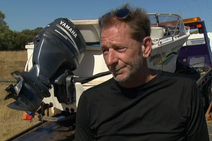 A man in a black top in front of a boat and trailer being loaded onto a tow truck.