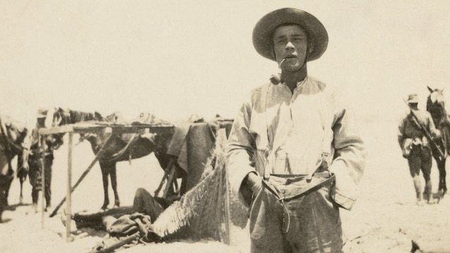 Photo of man smoking pipe with hands in his pockets and a horse in the background.