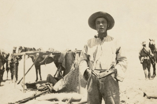 Photo of man smoking pipe with hands in his pockets and a horse in the background.