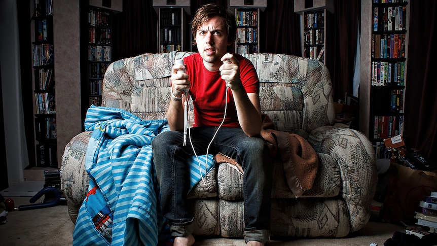 Man on couch holding gaming controllers, messy room