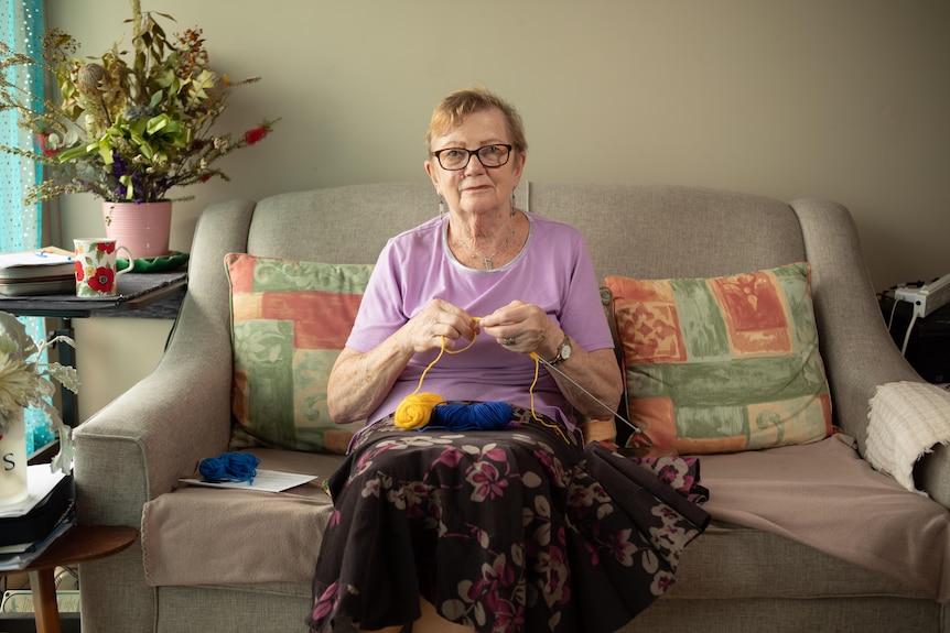 Olwyn-Ann sits on her couch holding her knitting.