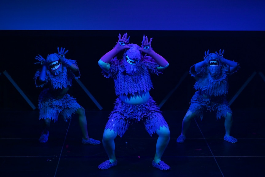 A darkly-lit stage photo of three people in dinosaur-like costumes with eyes on the dancers' hands