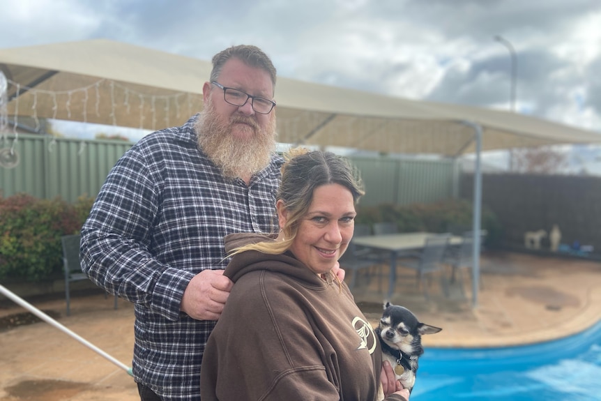 A middle aged couple hold a dog next to a swimming pool in a backyard