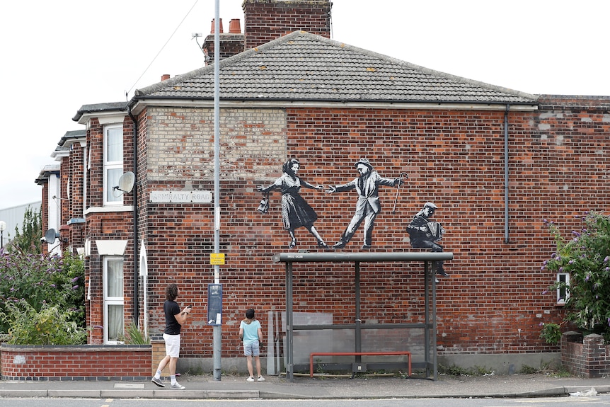 people look at a banksy artwork on a wall of people dancing above a bus shelter with a figure painted playing the acordian