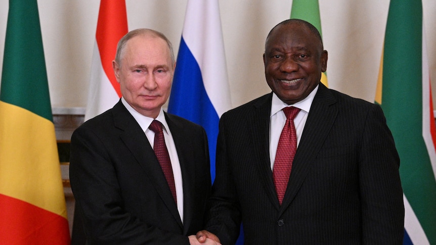 Putin shaking hands with Cyril Ramaphosa who is smiling infront of colourful flags. Both are wearing black suits and red ties.