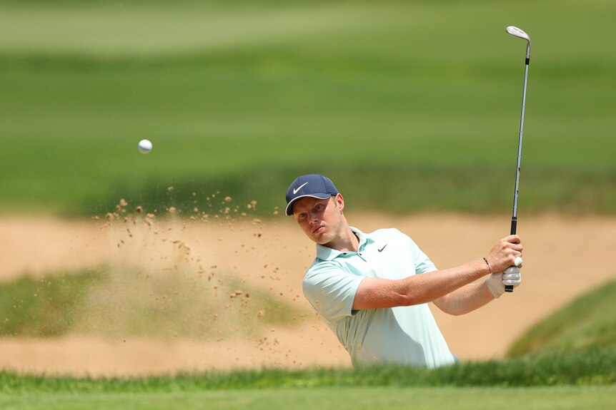A golfer stands in a bunker looking towards the hole as his shot sprays sand in the air.