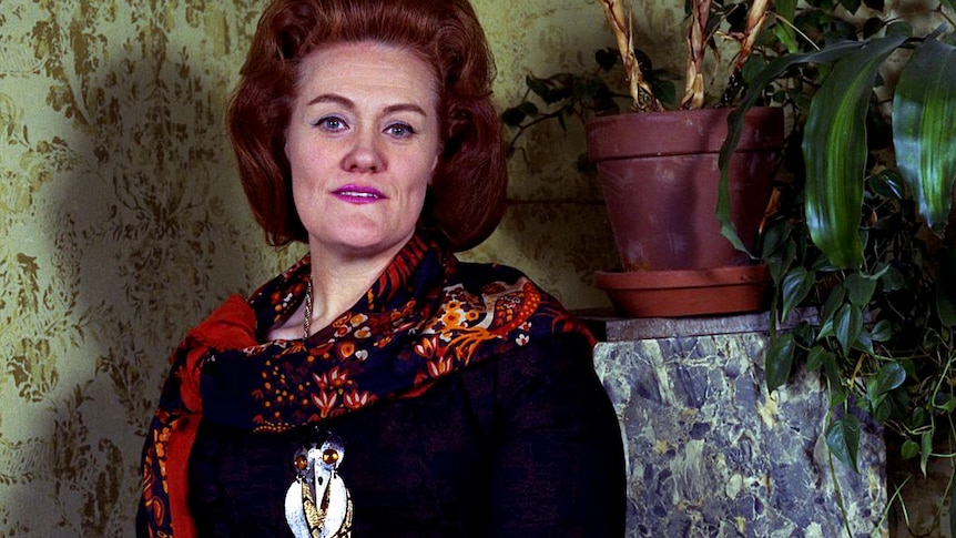 Dame Joan Sutherland poses in front of a wallpapered wall surrounded by plants.