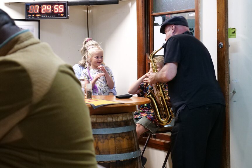 Mark McGurgan playing the saxaphone to a little girl with braids sitting in the pub on her mothers lap. 