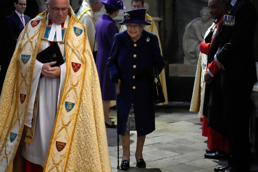 女王身着紫色衣服，拄着拐杖在威斯敏斯特教堂内行走。