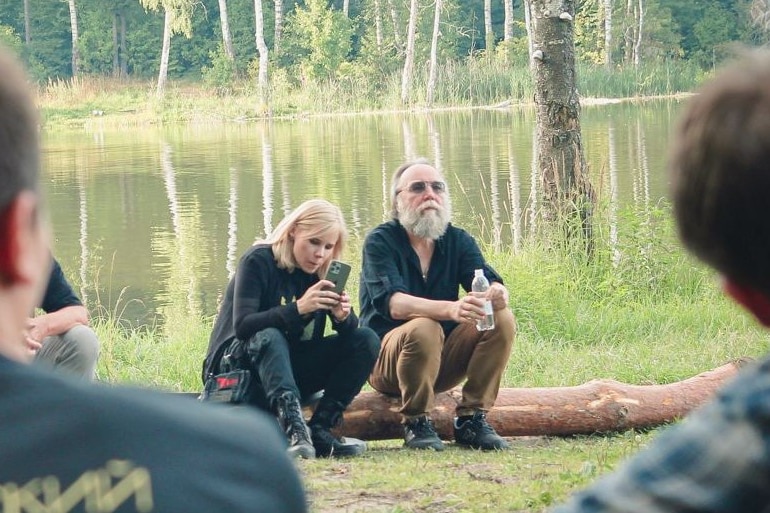 An older bearded man and a young blonde woman sit together on a log near a lake