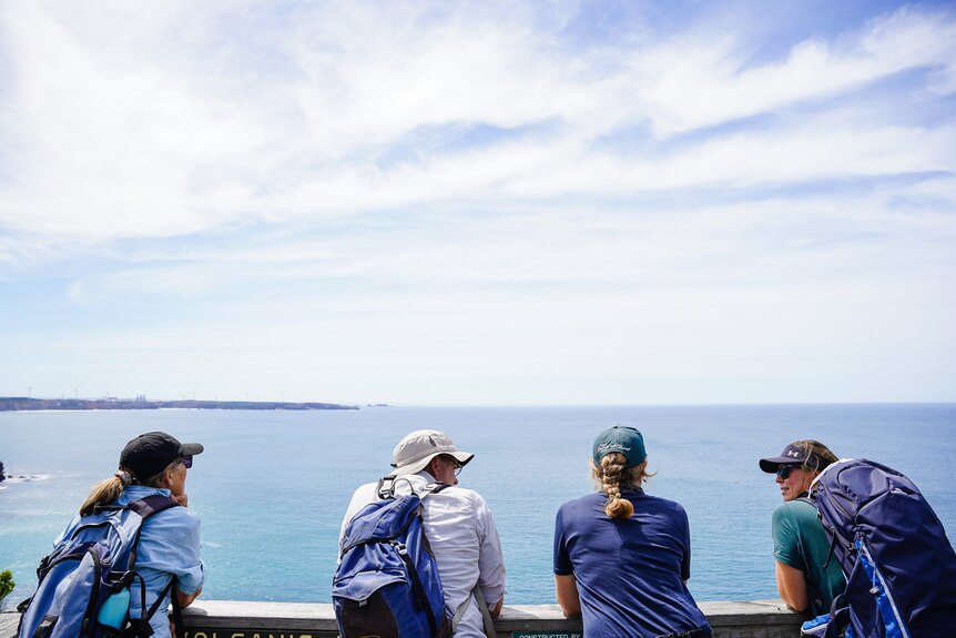 Four people in hiking gear lean against a wooden rail, looking out over the ocean under a clear blue sky.
