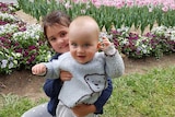 A little girl hugs a baby boy while standing on the grass with flowers behind them