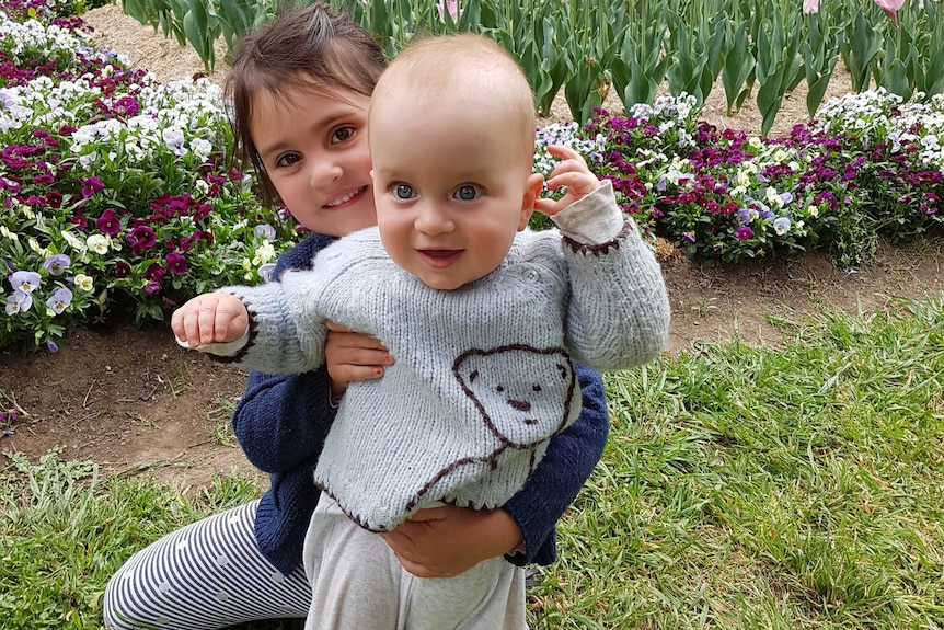 A little girl hugs a baby boy while standing on the grass with flowers behind them