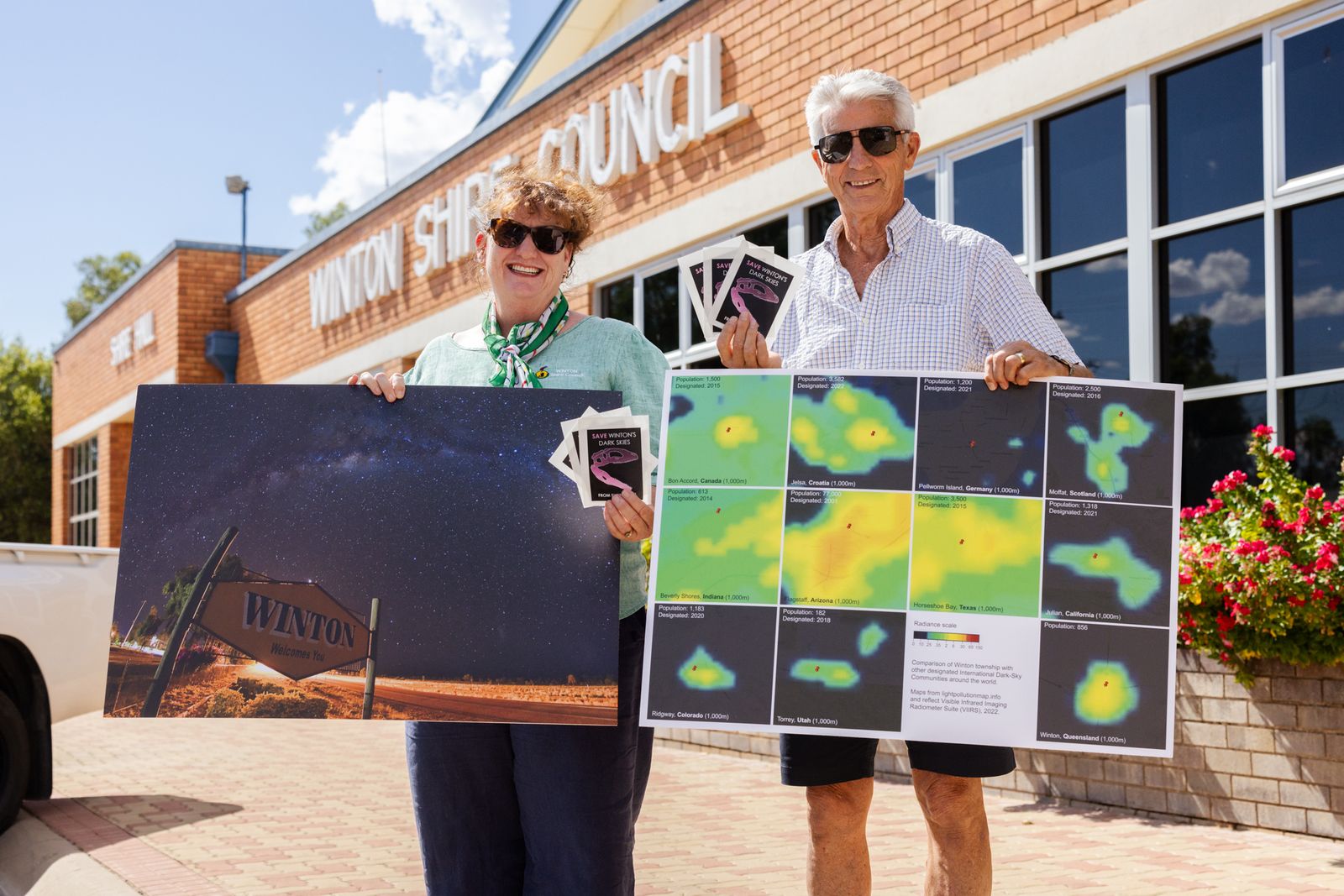 A smiling woman and man hold up large photographs in front of a brick council building.
