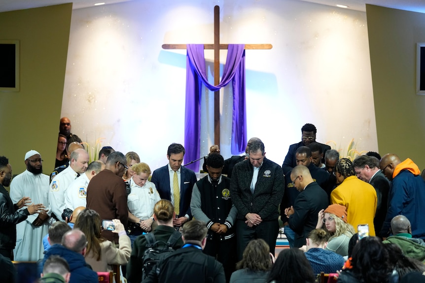 People stand with heads bowed in front of a large cross draped with purple fabric