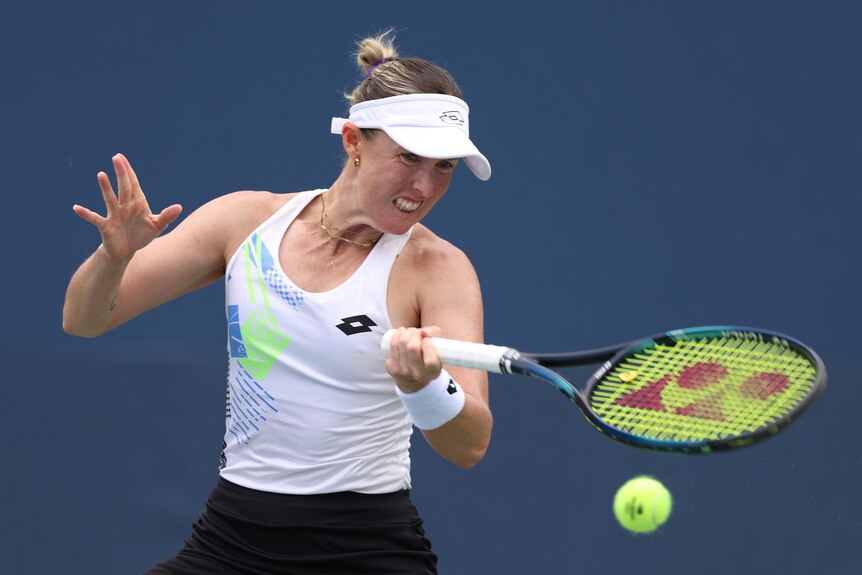 A female tennis player in a white visor, white top, grimaces she plays a forehand shot
