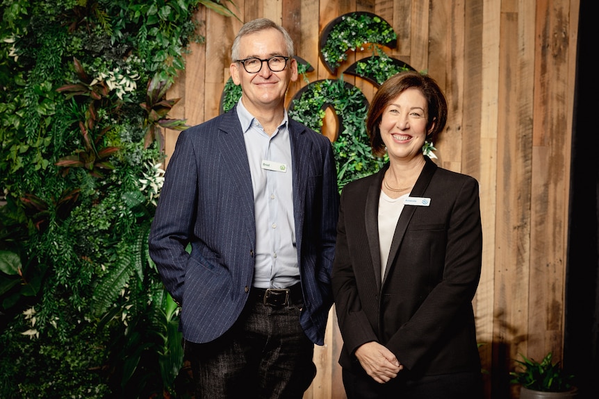 A middle-aged man and woman in suits stand smiling in front of a wooden backdrop with plants.