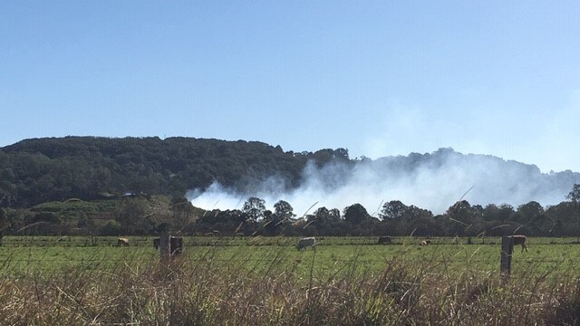 Smoke seen from a distance blows over paddocks and hills in a green area.