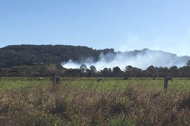 Smoke seen from a distance blows over paddocks and hills in a green area.