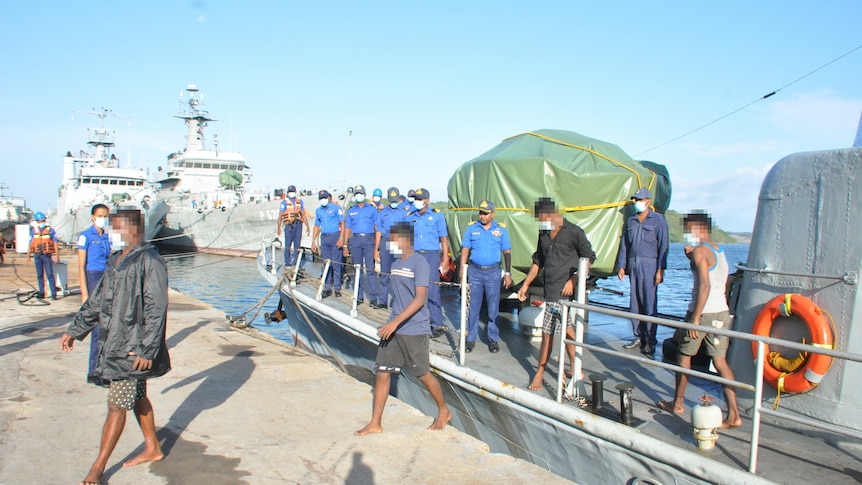 A group of people walk of a boat while they are watched by a group of men dressed in blue.