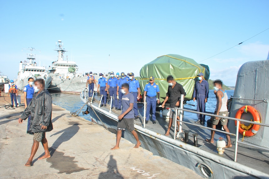 A group of people walk of a boat while they are watched by a group of men dressed in blue.