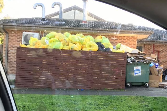 Yellow bags of medical waste pile high above a wooden rubbish enclosure outside a brick building.
