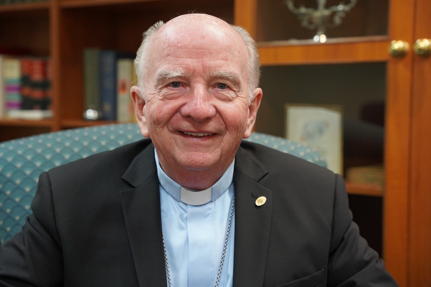 An older man sitting down. He is wearing a priest's collar and a black suit jacket.