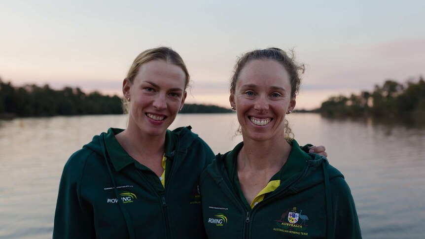 Two women have their arms around each other, wearing Rowing Australia green jumpers. River and trees blurred in the background.