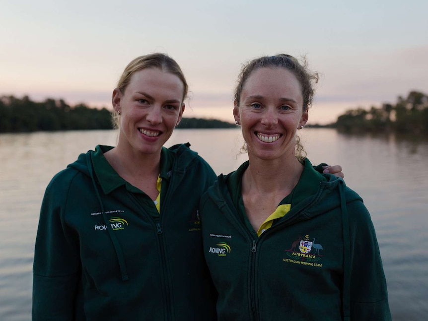 Two women have their arms around each other, wearing Rowing Australia green jumpers. River and trees blurred in the background.
