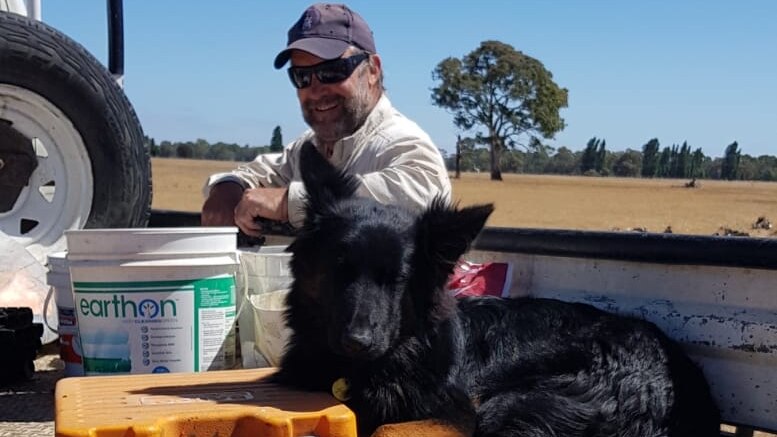 A farmer wearing a cap and sunglasses leaning onto his ute tray with a black dog sitting near him. 