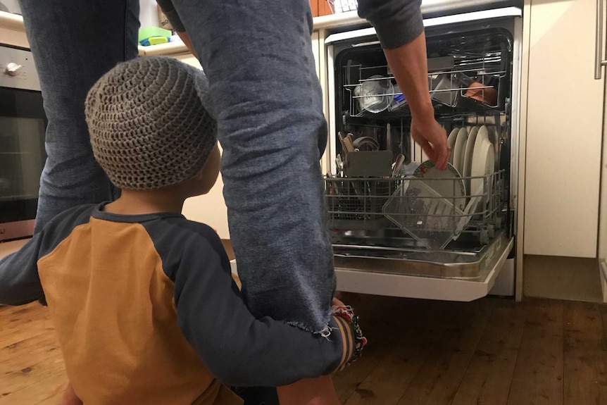 A person unpacks a dishwasher while a child hangs onto their leg for a story on parents feeling 'touched-out'.