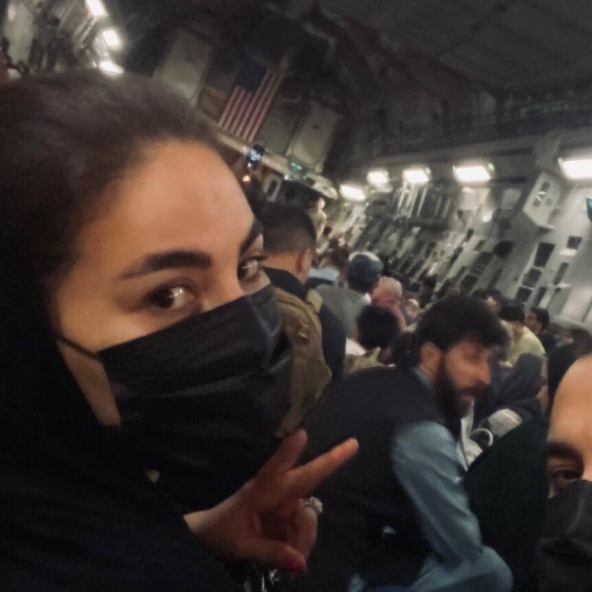 Aryana, wearing a face mask does a peace sign with her fingers in a crowded cargo hold of a military plane.