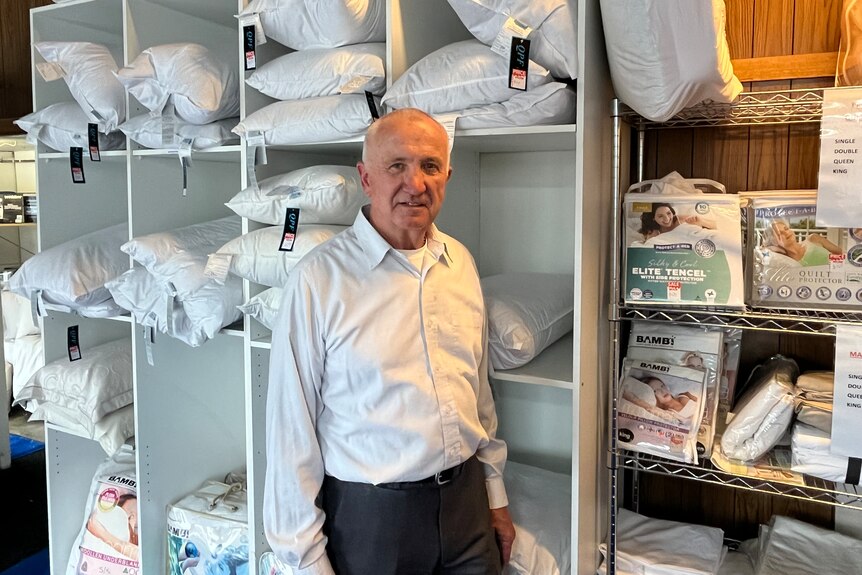 An older man wearing a collared shirt stands in front of shelves of pillows.