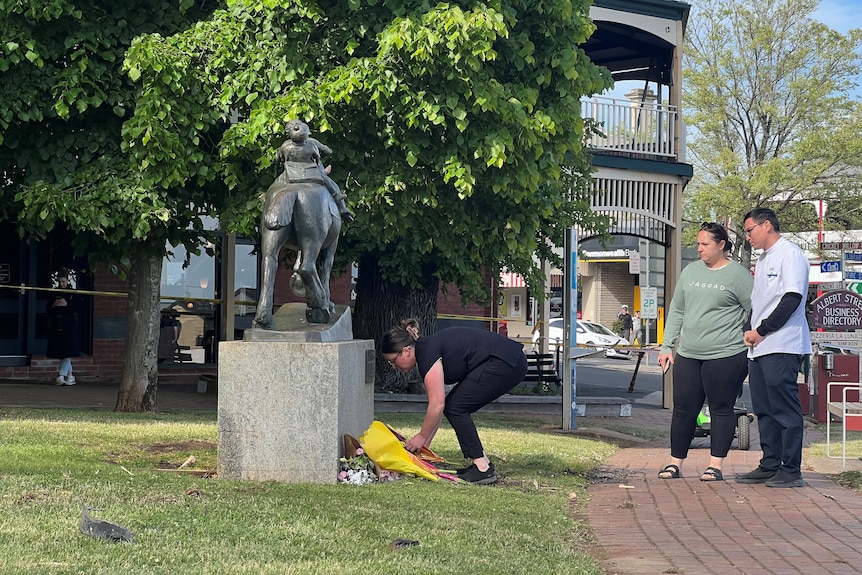 A woman bends down to place flowers at the base of a statue in a park while two other people look on.