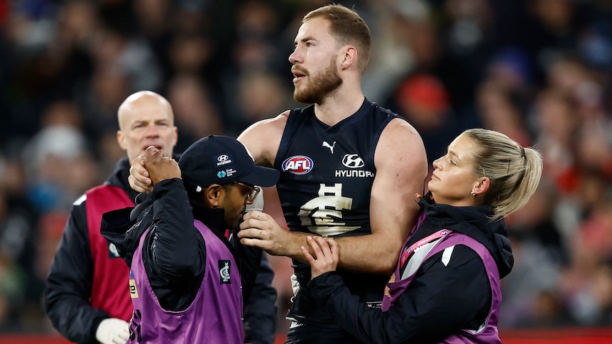 A Carlton AFL player is assisted from the field by trainers and medical staff.