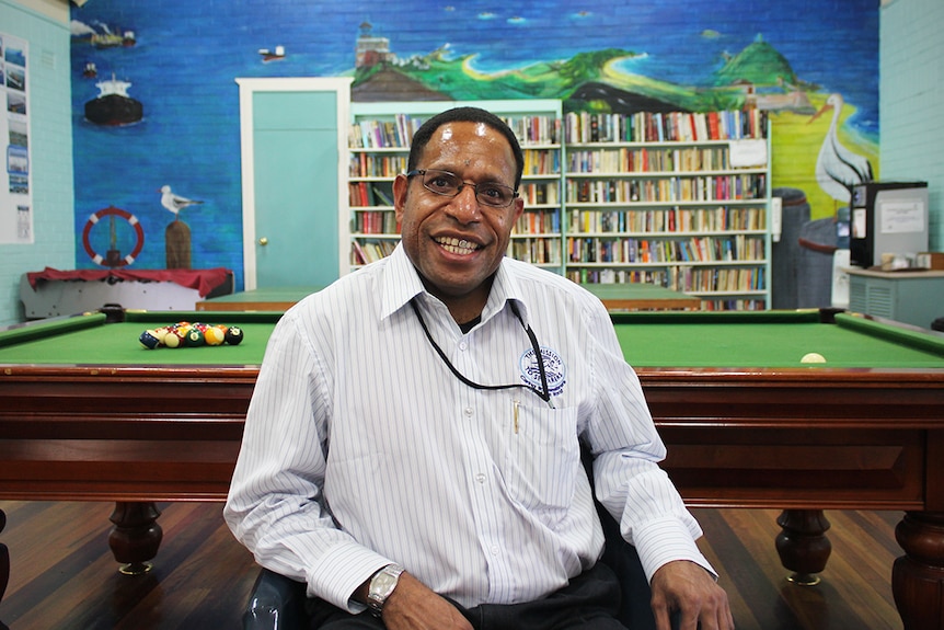 A smiling man sits in a chair in front of a pool table an ocean mural with a bookcase.