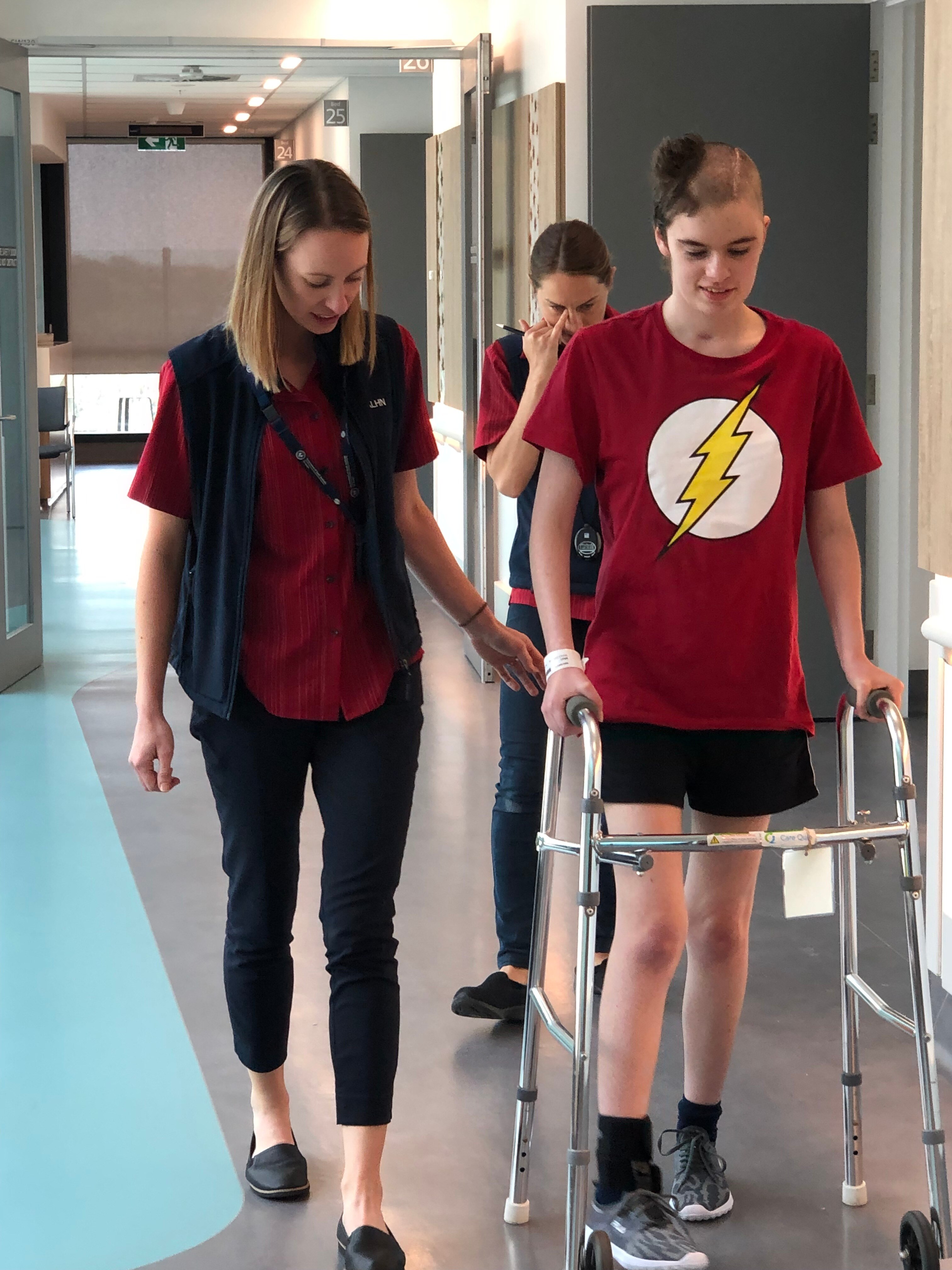 Nicola Bray uses a walker in a hospital corridor while flanked by hospital staff.