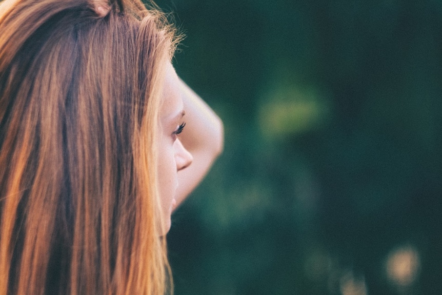 Woman with hand in light brown hair to the left looking into the right, where there is greenery blurred