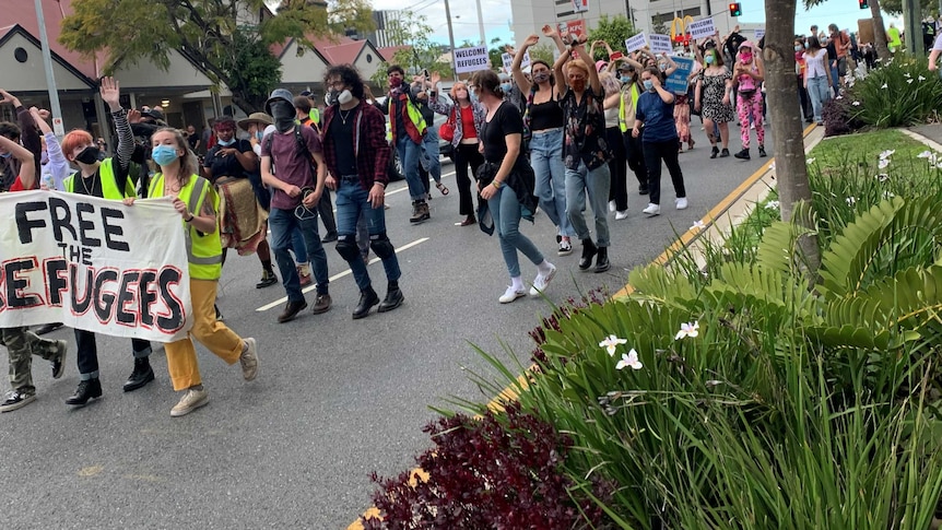 Hundreds of people, many wearing face masks, walk along a closed major road while holding protest placards.