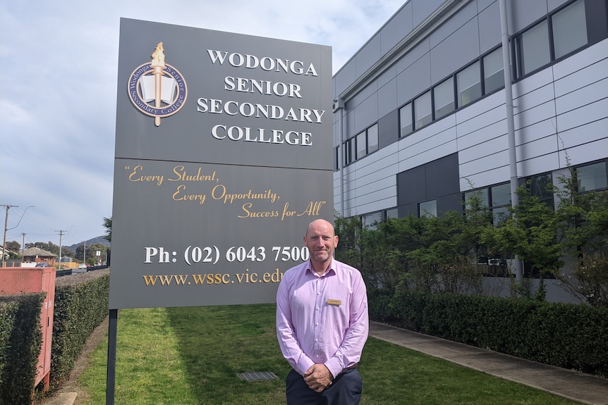 A man in a pink shirt and a name badge stands in front of the Wodonga Senior Secondary College school sign