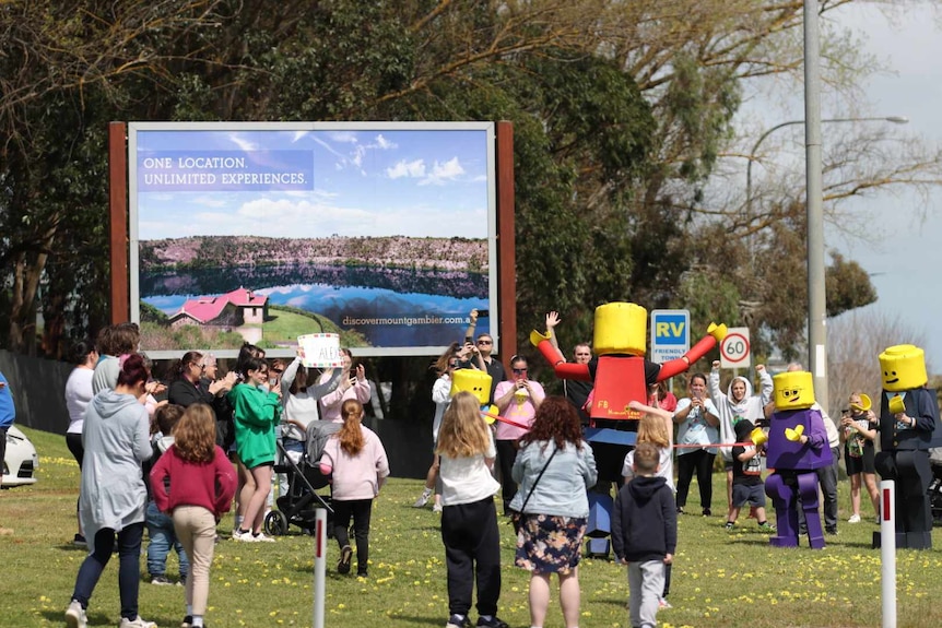 A man dressed as a Lego figurine is greeted by two others and people near a billboard.