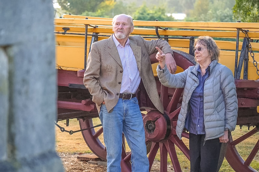 A man and woman lean against an old horse cart pointing to a large stone wall.