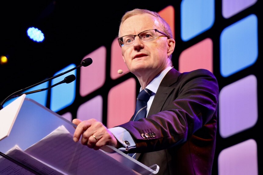 Philip Lowe stands at a lectern with a microphone. Behind him is a screen with multi-coloured squares