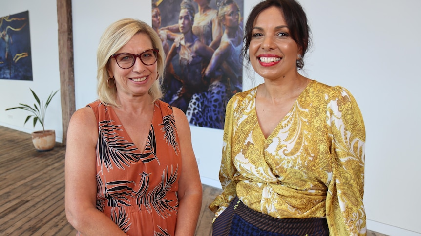 White woman with blonde hair wears orange top and dark glasses beside Kokatha woman with dark hair, gold shirt and red lipstick.